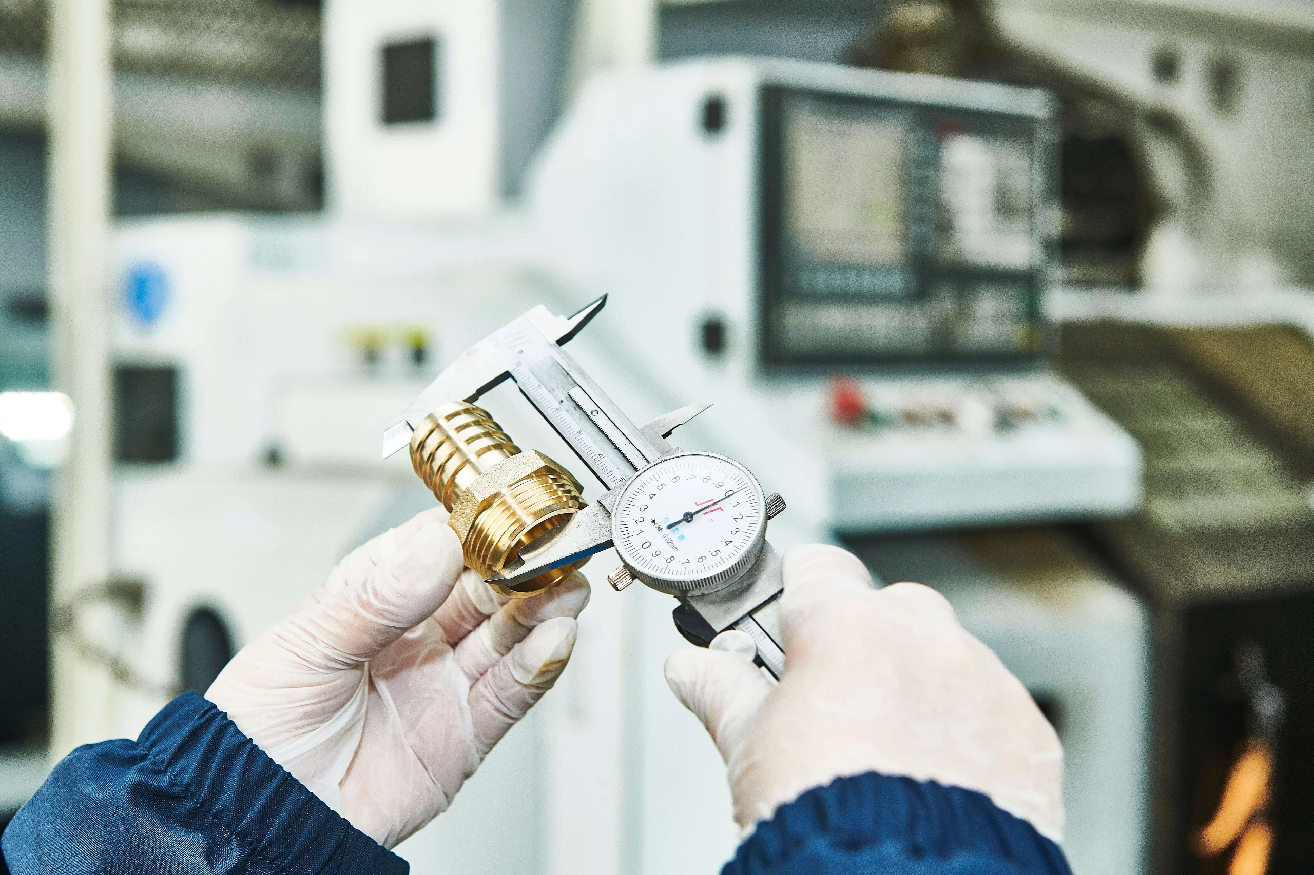 Quality control technician measuring a brass insert's dimensions with digital calipers on a factory inspection bench, next to a pull-out force testing rig