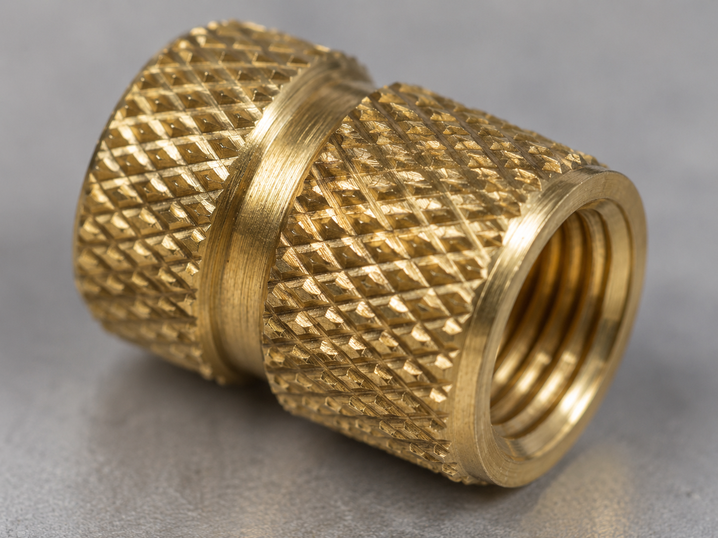 Close-up macro photo of a brass insert's knurled outer surface showing diamond-pattern grooves and a circumferential anti-rotation ring, before being molded into plastic